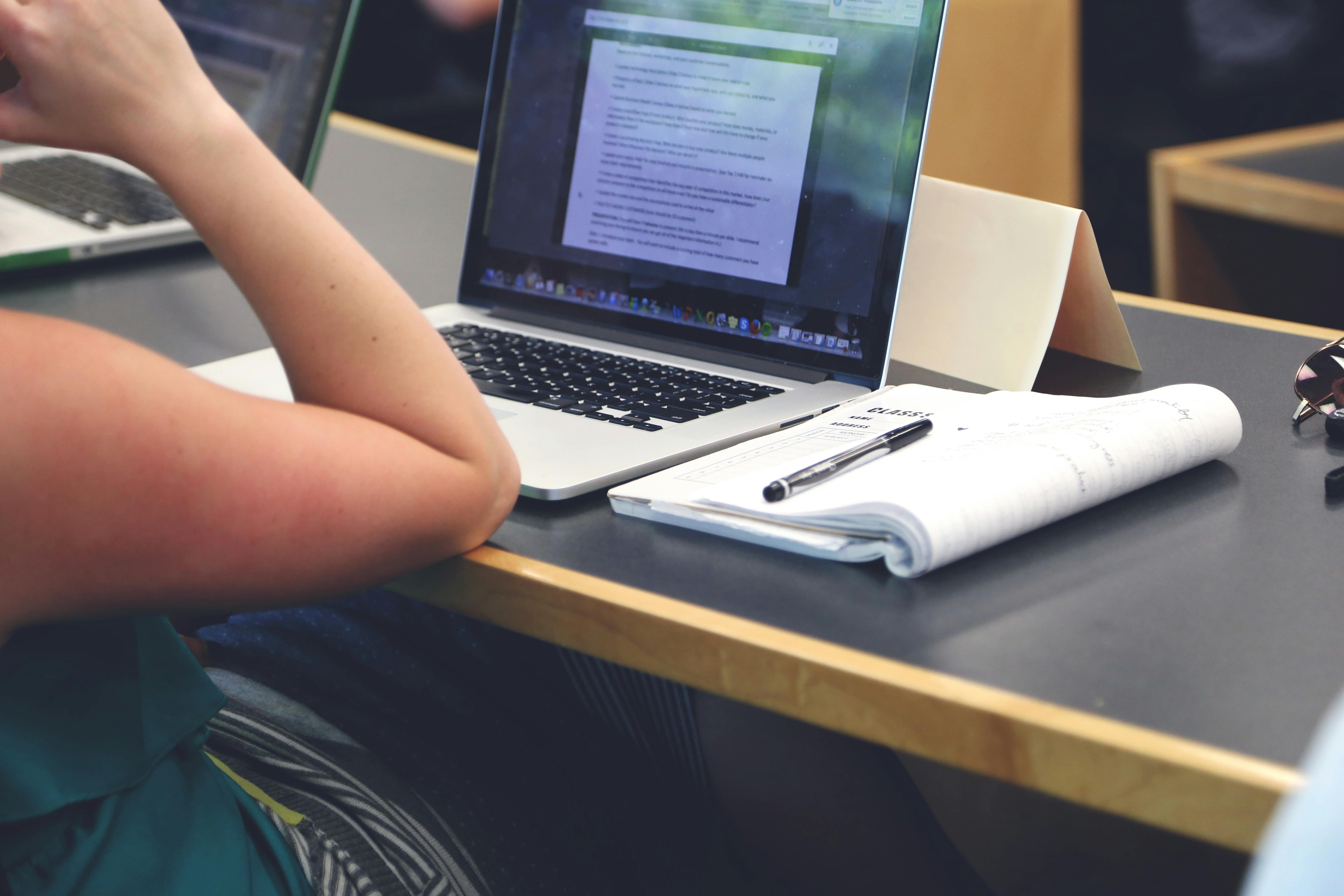 A student in a classroom using a laptop and taking notes in a notebook.