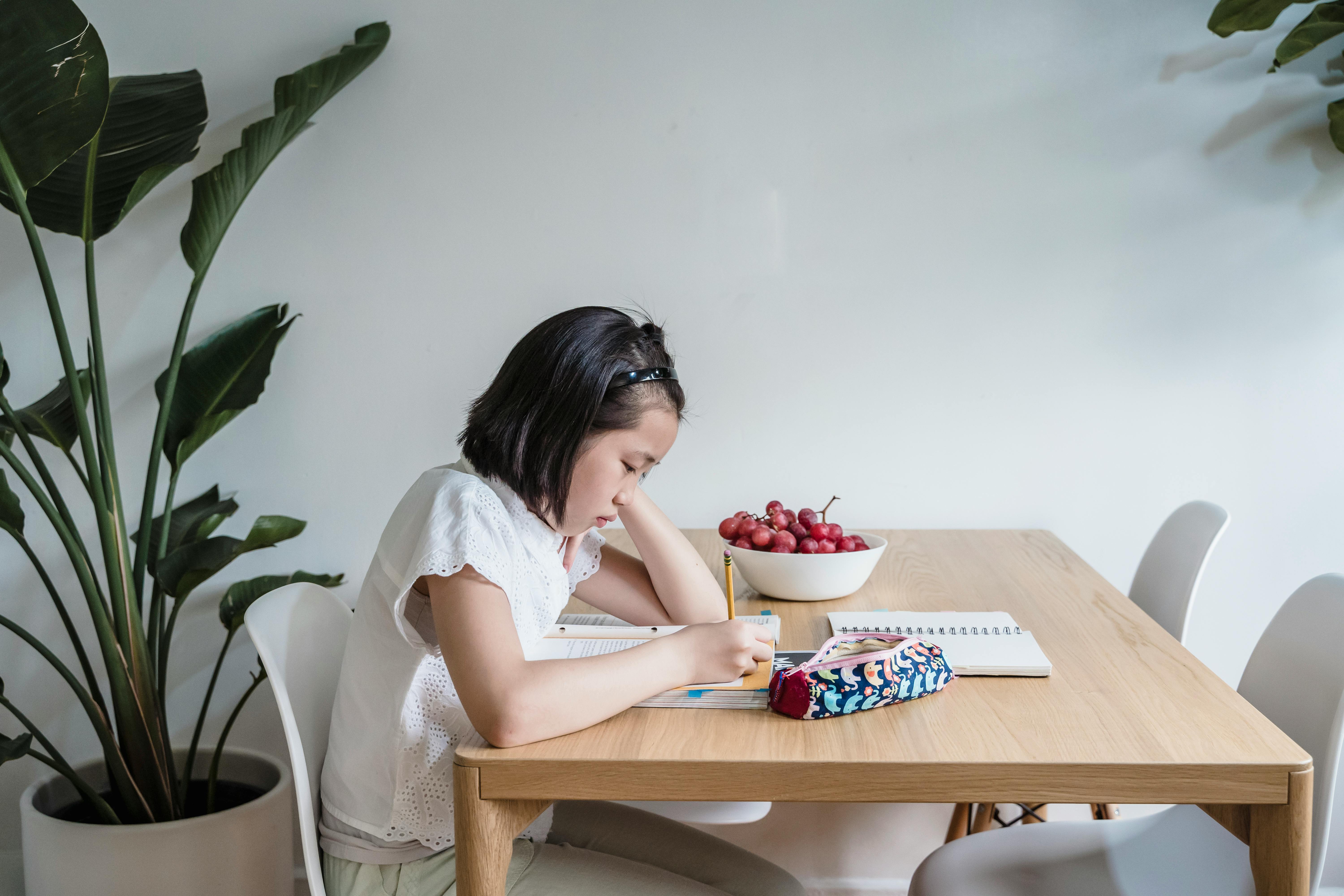 Asian child reading and writing at home, surrounded by plants and books, creating a learning-friendly environment.